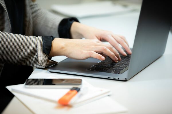 Das Bild zeigt ein Closeup von Händen, die an einem Laptop arbeiten. Daneben liegen Papiere, ein Textmarker und ein Smartphone.