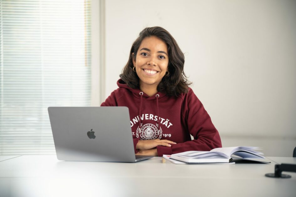 das Bild zeigt eine Studierende, die die Kamera anlächelt und mit einem Laptop und einem aufgeschlagenen Buch arbeitet. Es dient der Visualisierung der Arbeit mit Jupyter Notebooks.