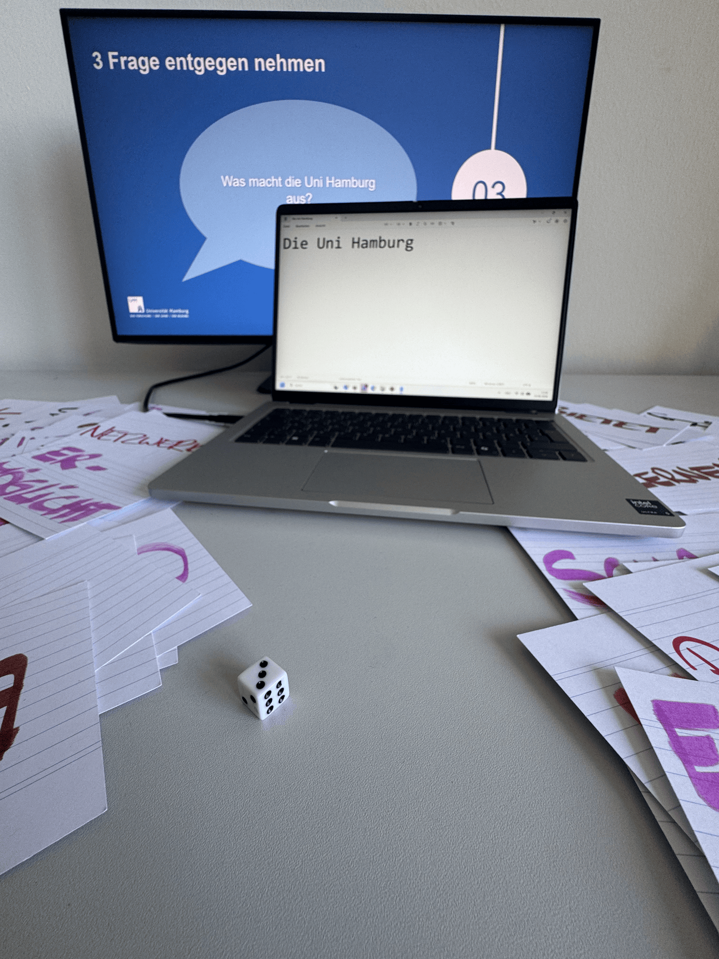 Ein GIF das zwei Bildschirme, Karteikarten und einen würfel zeigt. Auf dem hinteren Bildschirm steht due Frage "Was macht die Uni Hamburg aus?". Auf dem vorderen Bilschirm steht "Die Uni Hamburg". Mit jedem weiteren Foto im GIF verändert sich der Würfel und eine andere Karteikarte liegt vorne. Der Text auf dem vorderen Bildschirm wird mit den Karten weitergeschrieben. Im letzten Bild steht dort "Die Universität Hamburg ermöglicht Forschung, Lehre und Innovation."